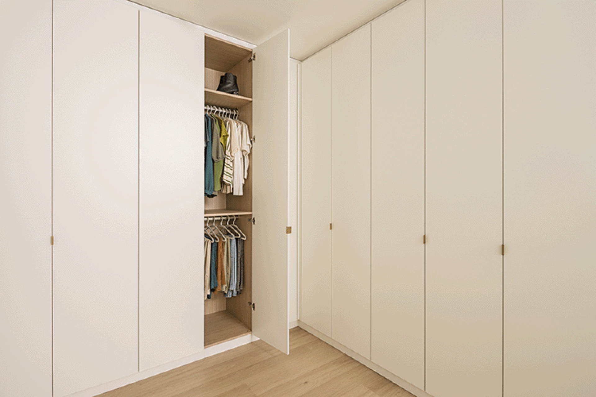 Foto van een L-vormige kledingkast op maat in Front White met messing Mini Pure handgrepen. De meest linkse deur die zichtbaar is op de foto werd geopend waardoor we 2 kledingroedes zien en een hoge legplank met kleding.