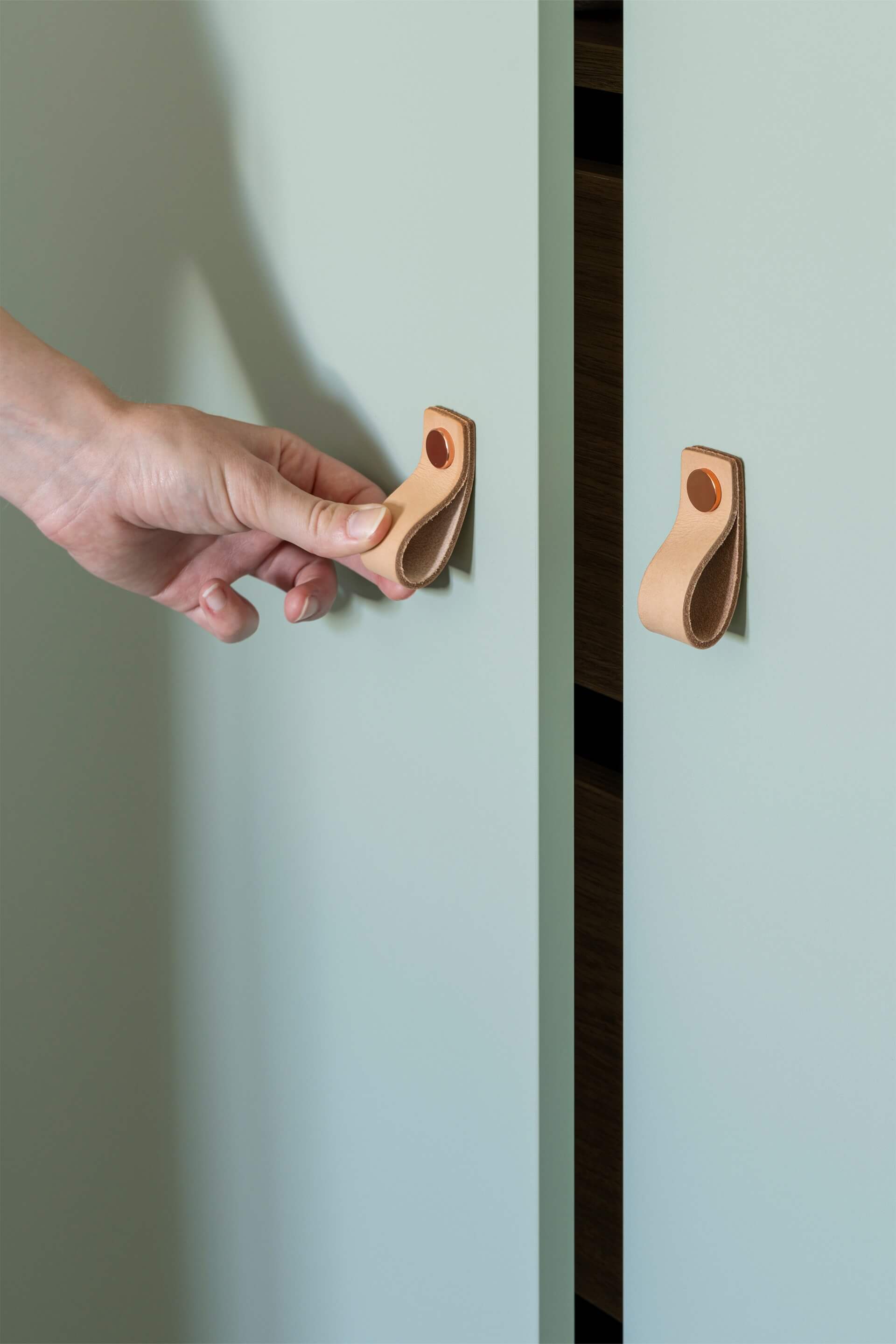 Gros plan sur un meuble sur mesure de couleur Industrial Green, avec des poignées Leather Loop en cuivré. À gauche, on aperçoit une main ouvrant une porte à l'aide d'une poignée.